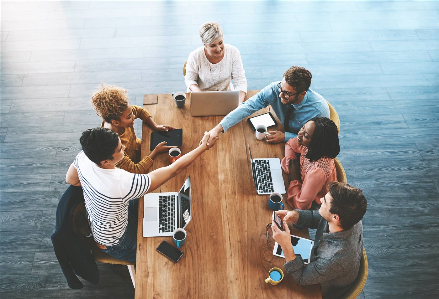 Overhead view of six people at a boardroom table; two reaching across to shake hands over laptops, tablets and documents.