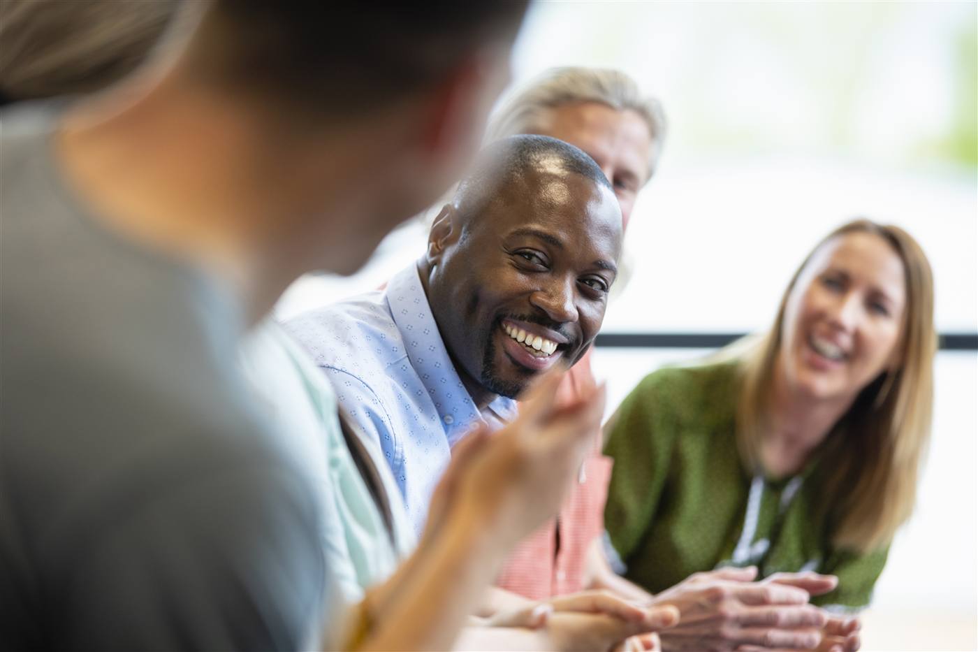 Three professionals in a workplace meeting: smiling bearded man foreground, woman in a green cardigan, older man behind.