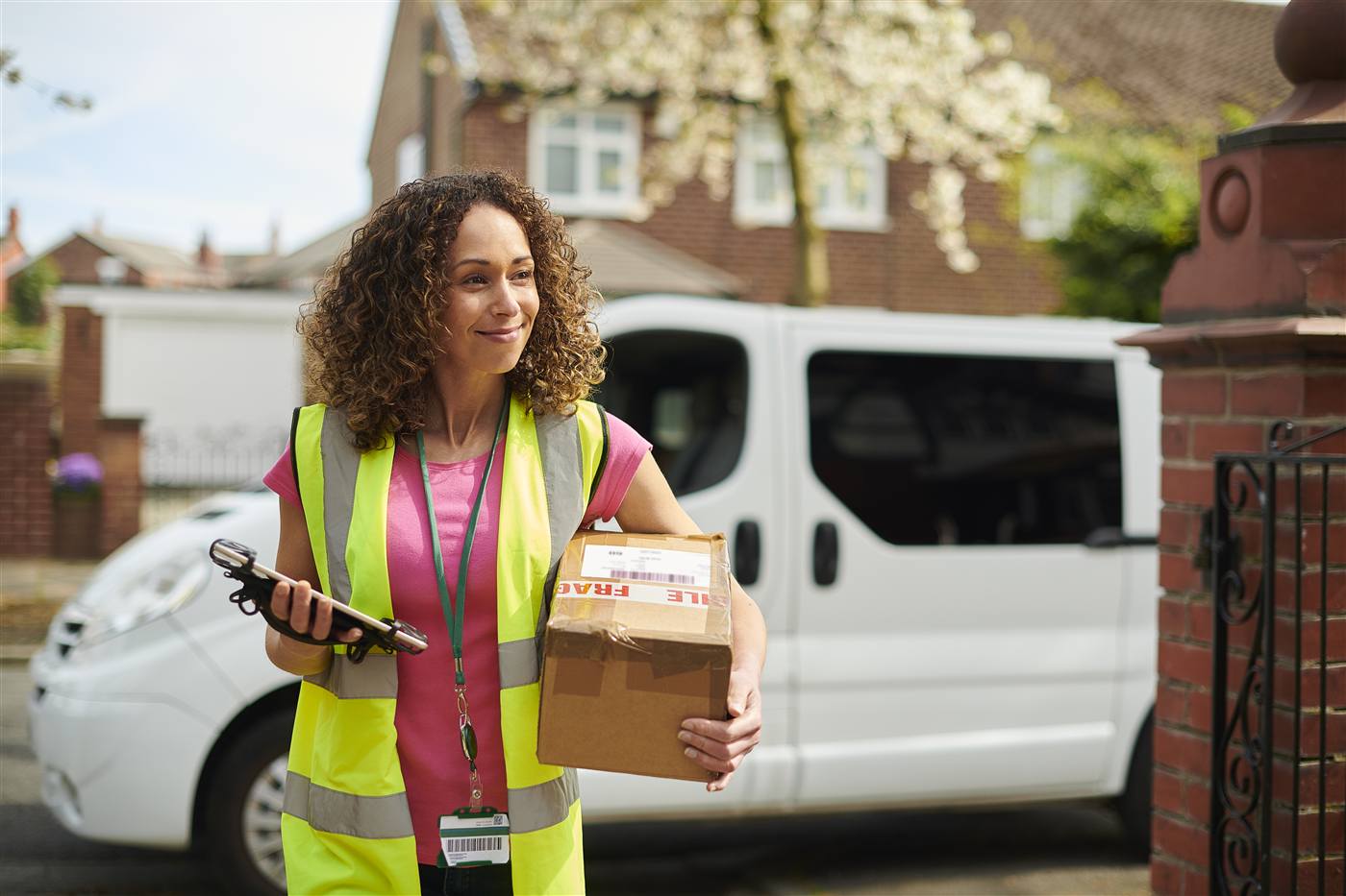 Female courier in a yellow hi-vis vest holds a parcel and barcode scanner at a residential gate; delivery van behind her.