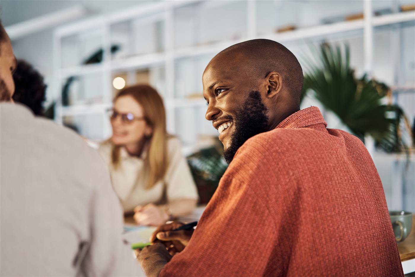 Smiling bald Black man in an orange-red shirt turned towards a colleague in a bright open-plan office.