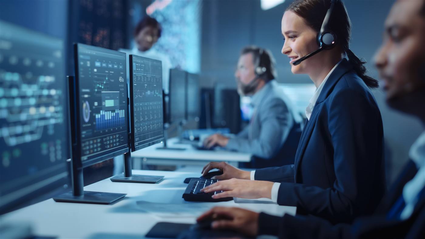Woman in navy jacket and headset at a workstation in a dimly lit operations centre with large data dashboard screens.