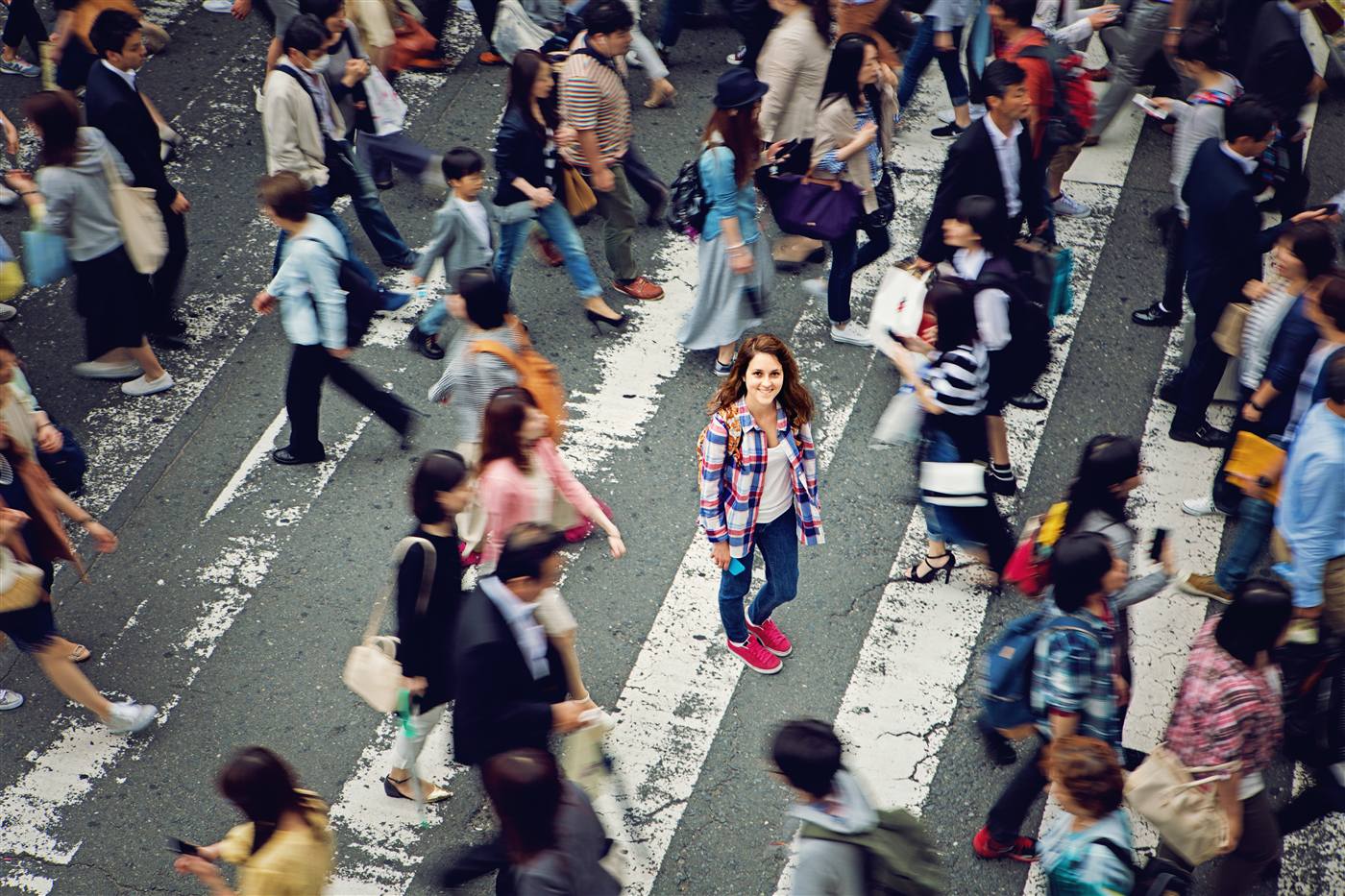 Woman with red hair in a check shirt stands still at a busy pedestrian crossing, surrounded by motion-blurred commuters.