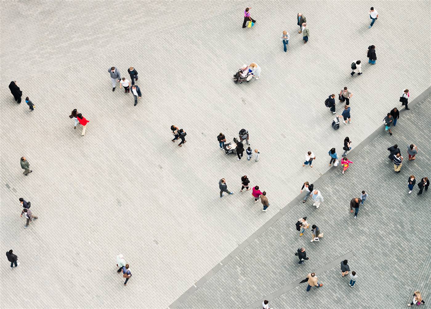 Aerial view straight down onto a grey plaza with dozens of tiny pedestrians casting short shadows, walking in all directions.