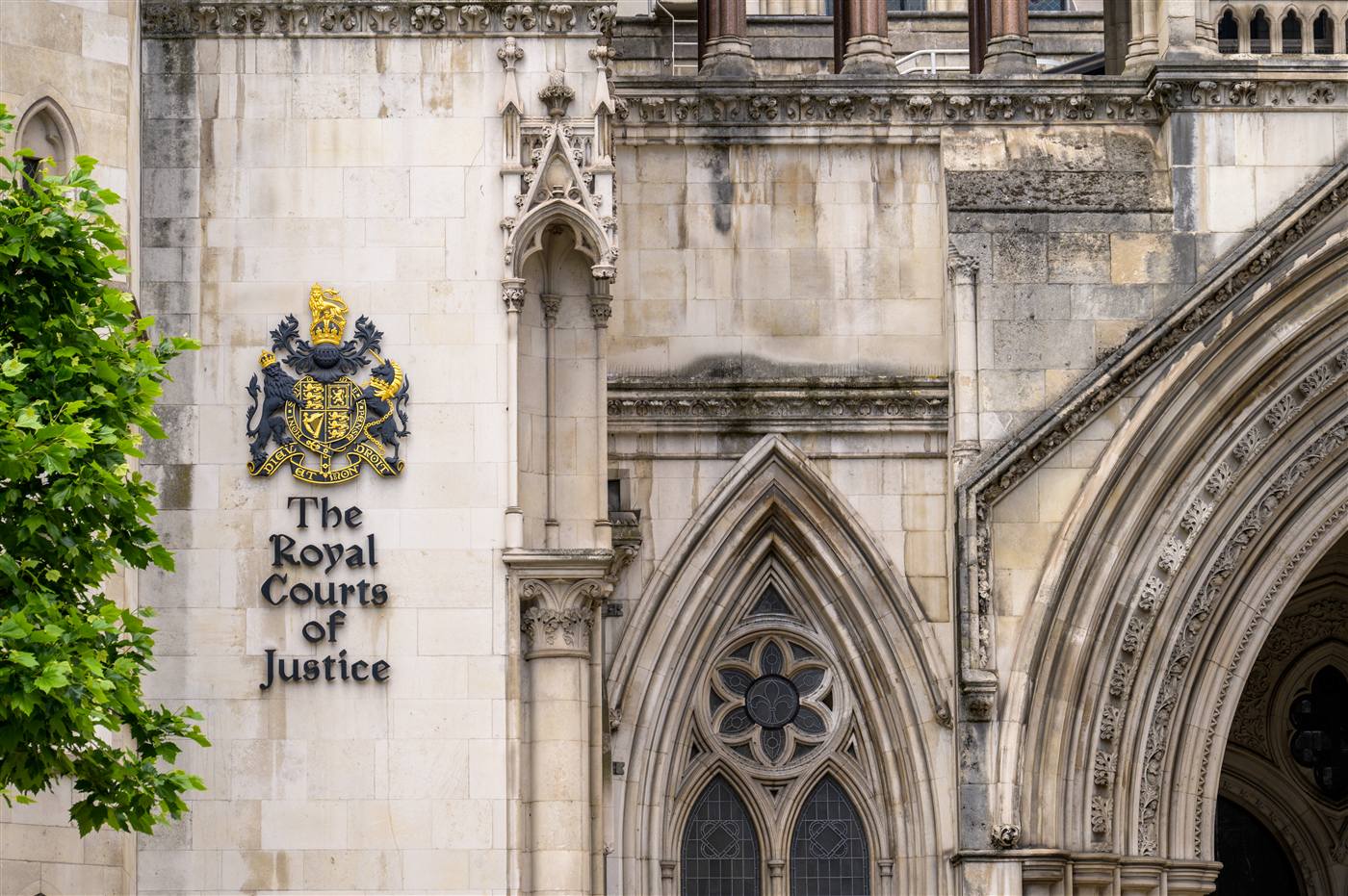 Exterior facade of the Royal Courts of Justice in London: pale stone neo-Gothic arched windows and a gold Royal Crest plaque.