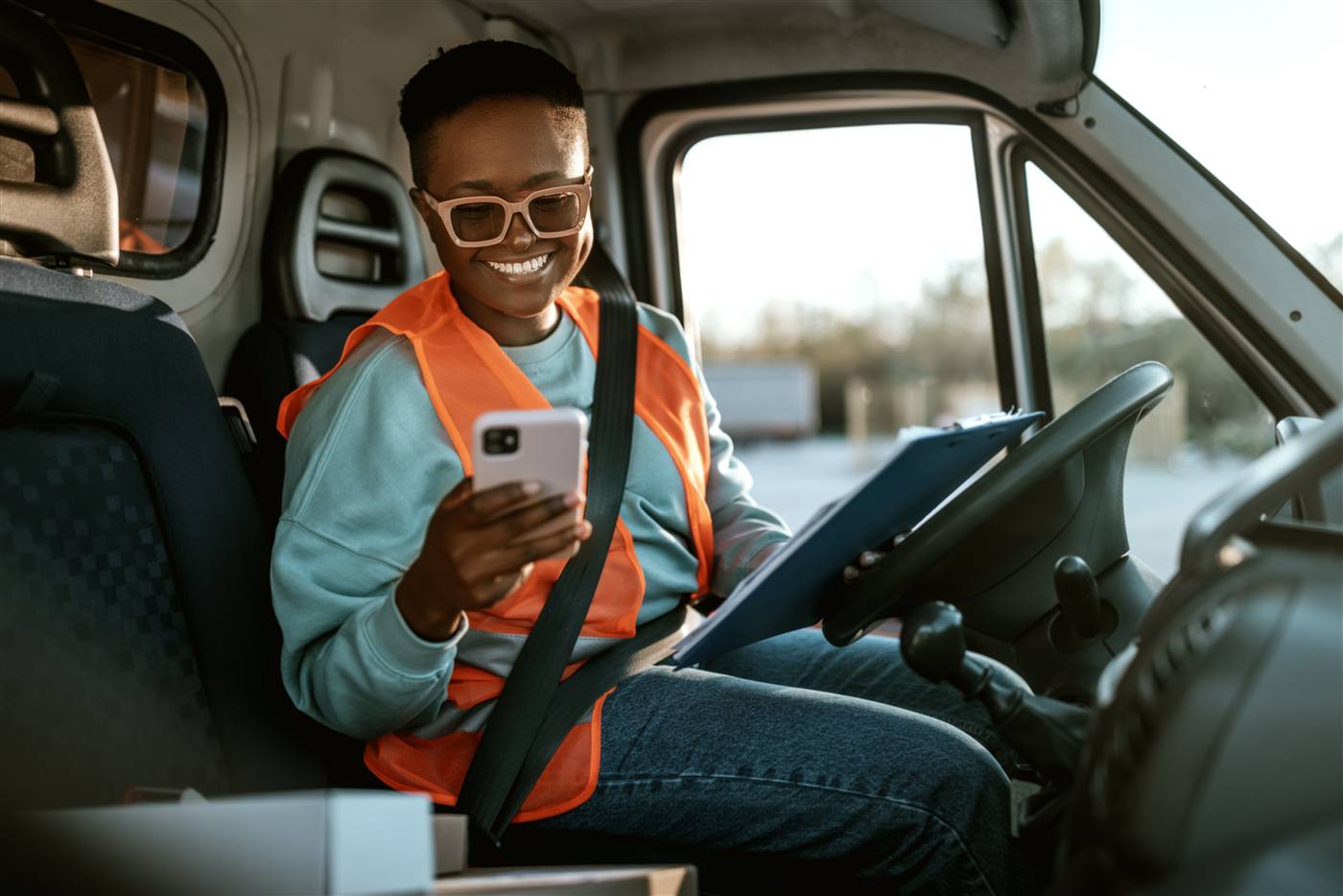 Smiling young Black woman in an orange hi-vis vest seated in a delivery van cab, holding a smartphone and a clipboard.