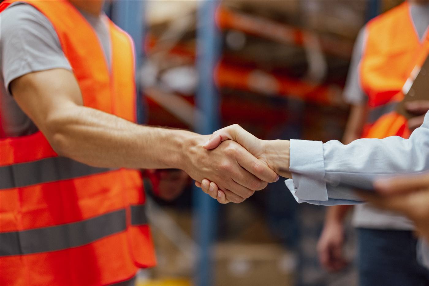 Close-up handshake between a hi-vis warehouse worker and a businessperson holding a tablet, with pallet racking behind them.