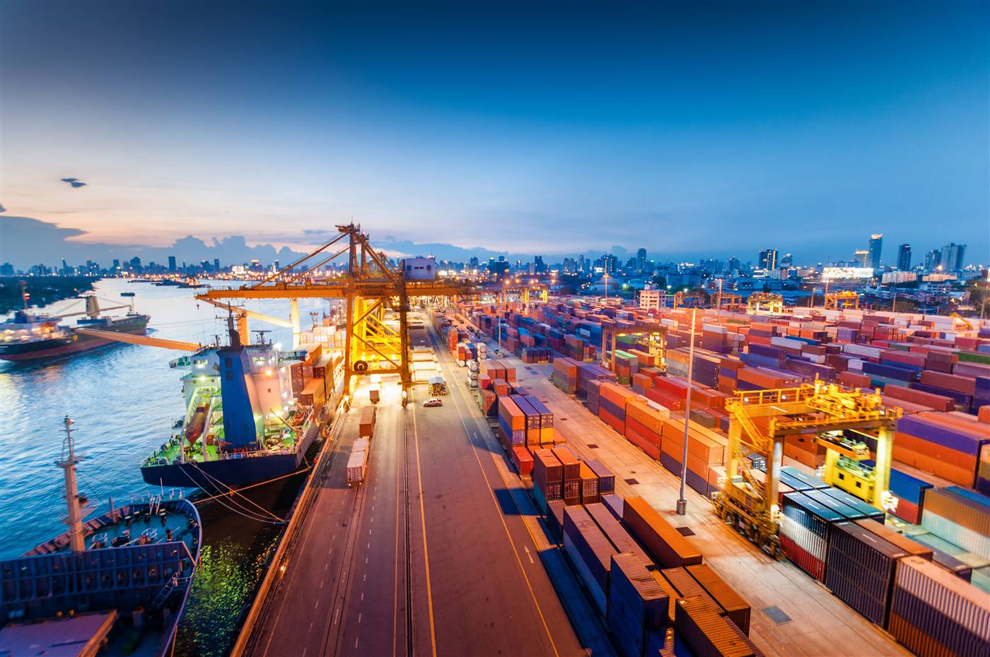Aerial dusk view of a shipping port with stacked containers, two yellow cranes, a moored cargo vessel and a harbour estuary.