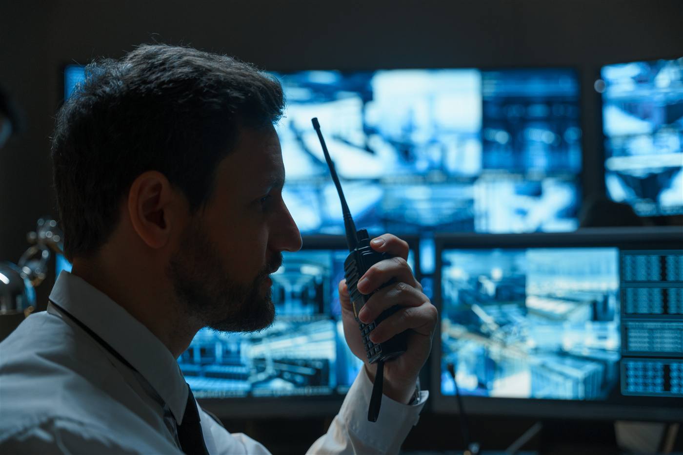 Man in a white shirt holding a walkie-talkie in a darkened room, lit by a bank of multiple CCTV and data screens behind him.