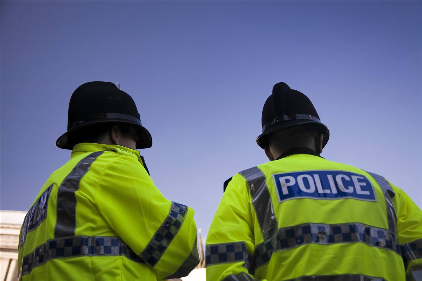 Two UK police officers from behind in yellow high-visibility jackets and black custodian helmets against a clear blue sky.